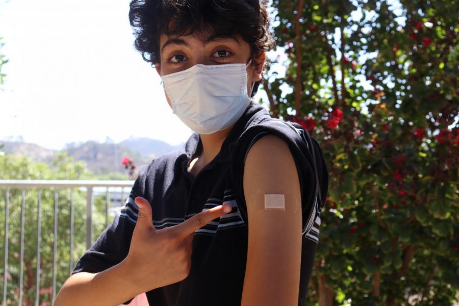 A student stands outside pointing towards a band-aid on their arm to show that they have their COVID-19 vaccine.